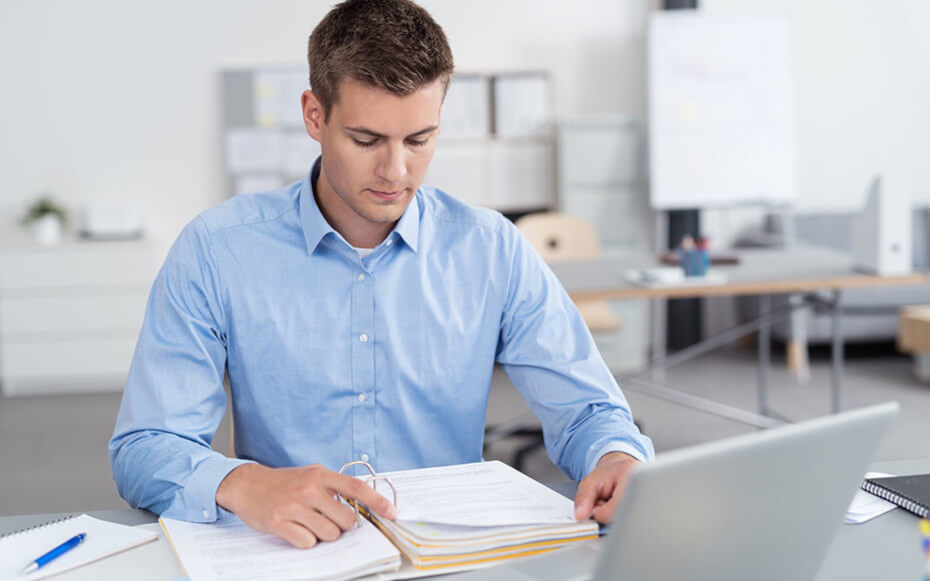 man sitting at desk reading a binder full of documents